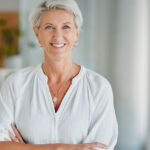 Dekorativt. Hvid kvinde med hvidt hår og hvid skjorte står med armene over kors. Hun smiler.