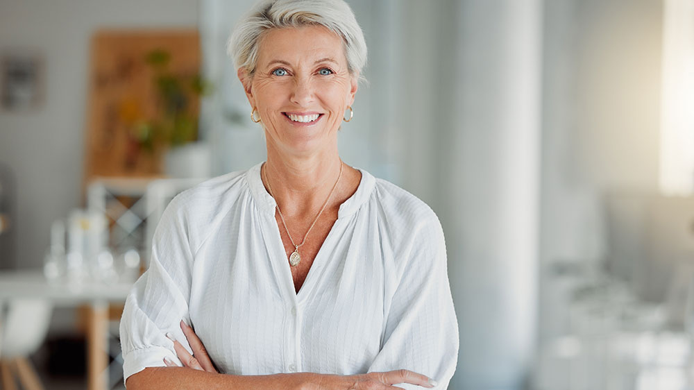 Dekorativt. Hvid kvinde med hvidt hår og hvid skjorte står med armene over kors. Hun smiler.